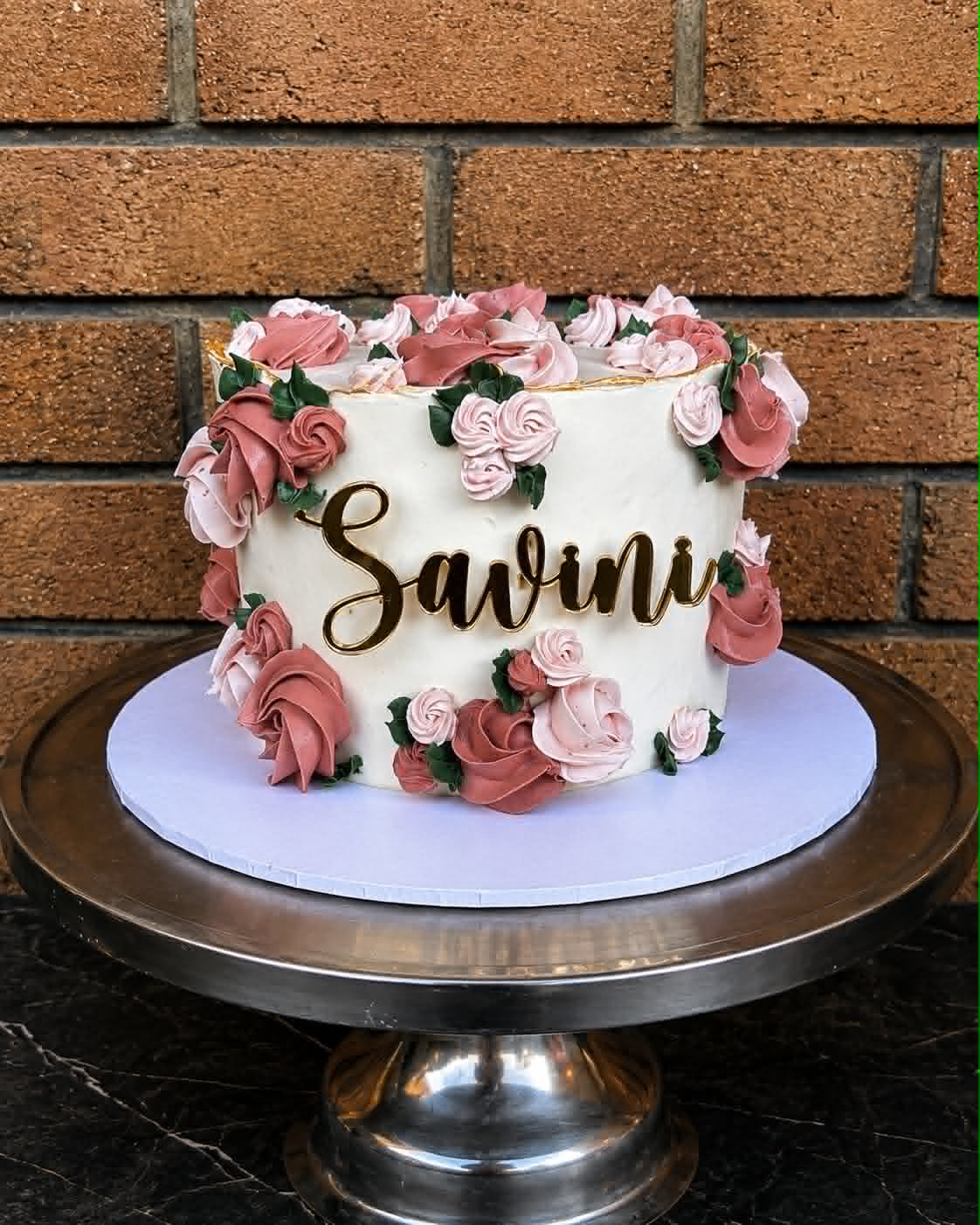White birthday cake with pink and dusty rose buttercream roses and gold acrylic name topper reading Savini